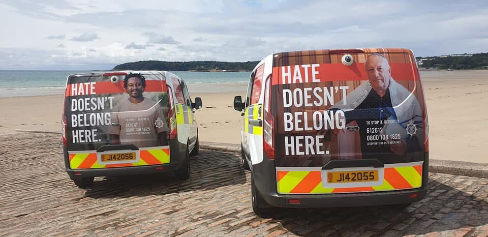 Two States of Jersey Police campaign vans parked on a Jersey beach, each wrapped with the 'Hate Doesn't Belong Here' campaign graphic featuring Martin Aliga and another islander alongside the 24/7 hate crime helpline number